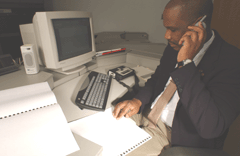 Image of a man reading braille while talking on a phone. A computer and braille notetaker are nearby